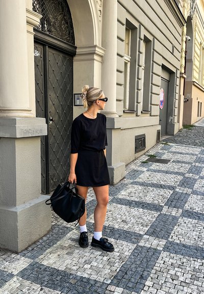 Vestido negro de manga corta combinado con zapatos negros, calcetines blancos y un gran bolso negro. De pie en una calle empedrada con un patrón.