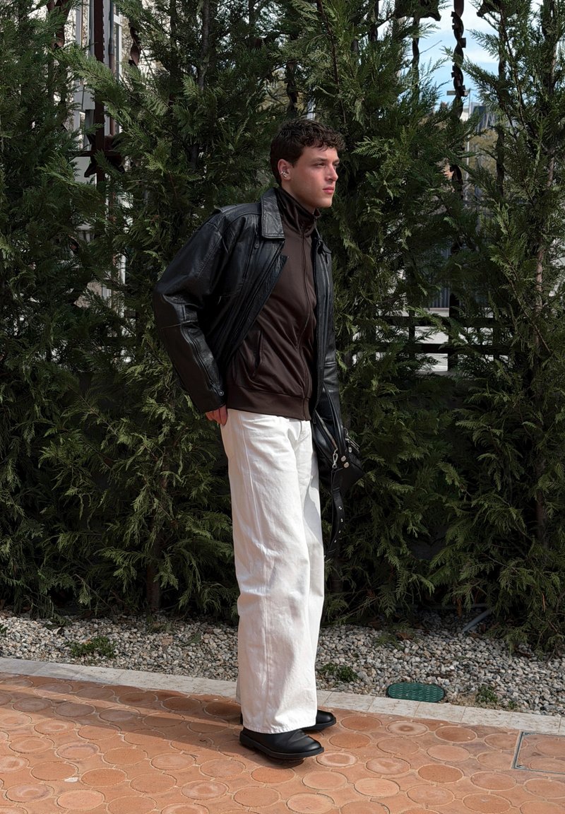 Jeune homme portant une veste en cuir noire, un haut marron à fermeture éclair, un pantalon blanc et des chaussures noires, debout sur un chemin carrelé avec des buissons verts derrière lui.