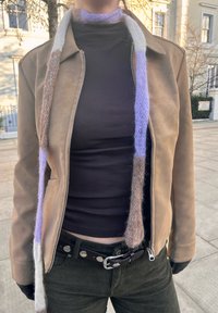 Veste en cuir marron avec une fermeture éclair, col roulé noir, pantalon en velours côtelé vert, et une écharpe tricotée multicolore avec des tons violets et marrons.