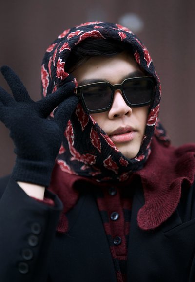 Gafas de sol negras, bufanda negra y roja con estampado, guantes negros y un abrigo oscuro con una camisa roja de botones. Vista en primer plano de la cara y las manos.