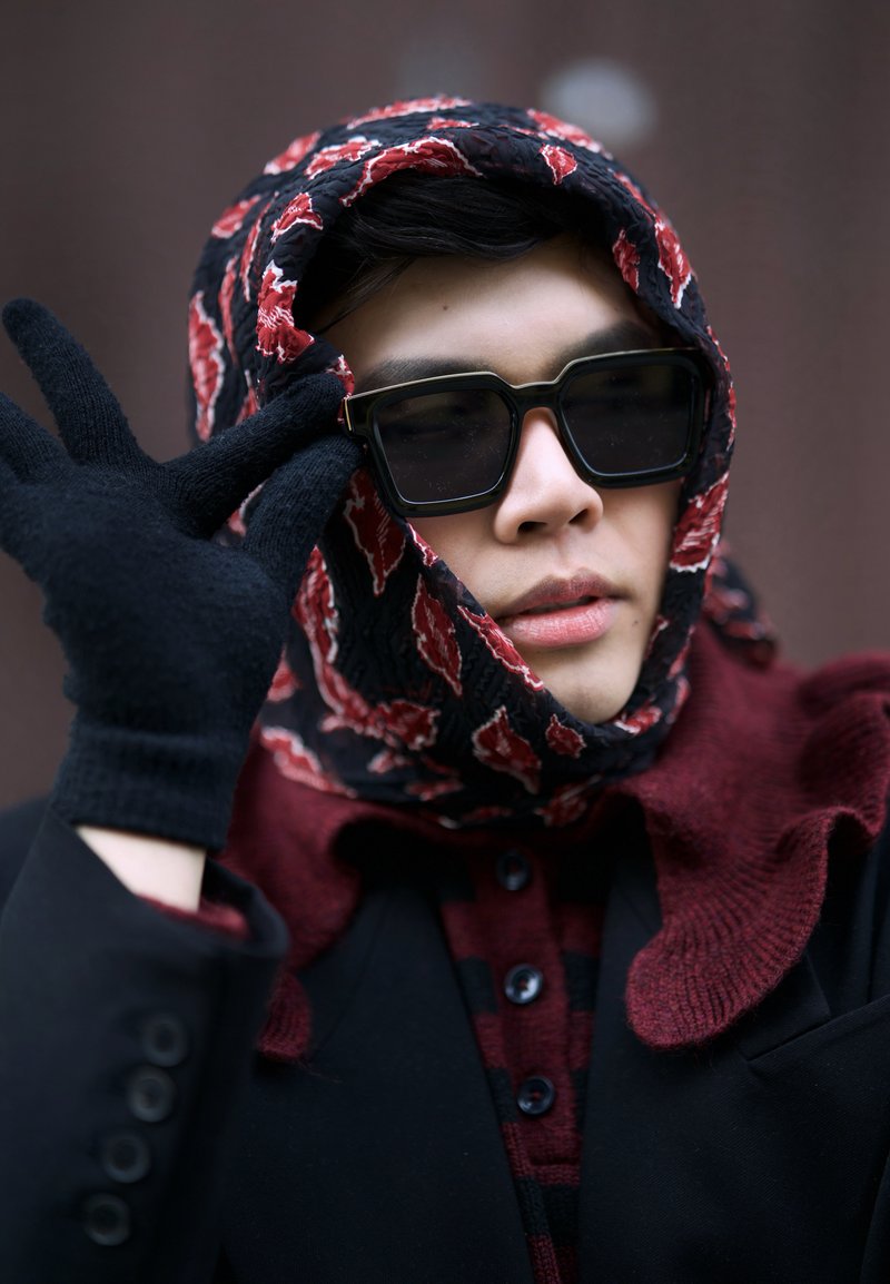 Nero occhiali da sole, sciarpa a motivi nera e rossa, guanti neri e un cappotto scuro con una camicia rossa abbottonata. Vista ravvicinata del viso e delle mani.