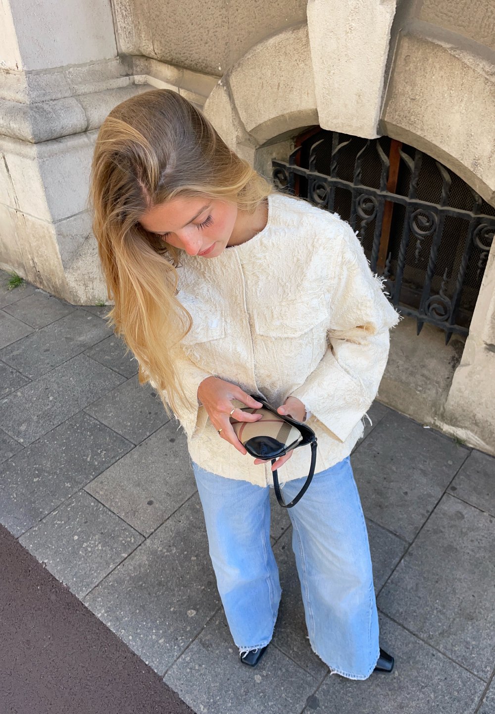 Casaco de creme texturizado com mangas oversized, combinado com jeans de perna larga azul claro. Segura óculos de sol pretos, em pé sobre uma superfície de azulejos cinzentos.