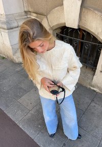 Casaco de creme texturizado com mangas oversized, combinado com jeans de perna larga azul claro. Segura óculos de sol pretos, em pé sobre uma superfície de azulejos cinzentos.