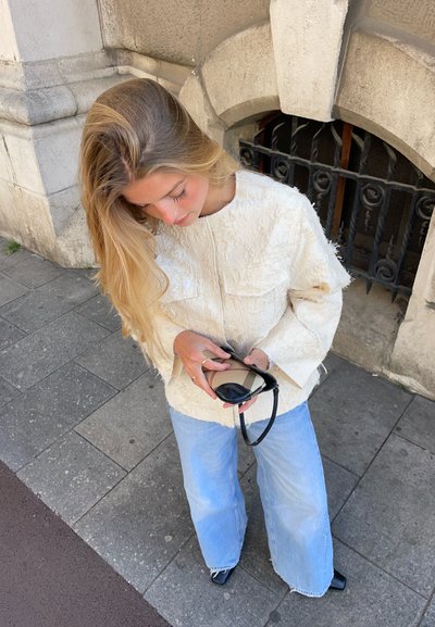 Chaqueta de crema texturizada con mangas oversized, combinada con jeans de pierna ancha de color azul claro. Sostiene gafas de sol negras, de pie sobre una superficie de baldosas grises.