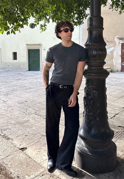 Camiseta ajustada de manga corta gris, pantalones de pierna ancha negros con rayas, zapatos negros y gafas de sol negras. De pie junto a un farol decorativo.