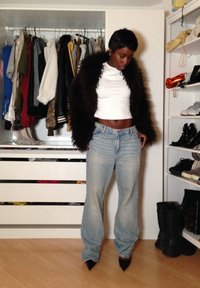 Personne portant une veste en fourrure noire, un crop top blanc, un jean bleu ample et des chaussures noires pointues, debout dans une pièce avec des vêtements et des chaussures.