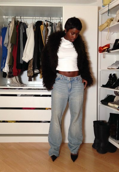 Persona con chaqueta de piel negra, top corto blanco, vaqueros azules holgados y zapatos negros puntiagudos, de pie en una habitación con ropa y zapatos.