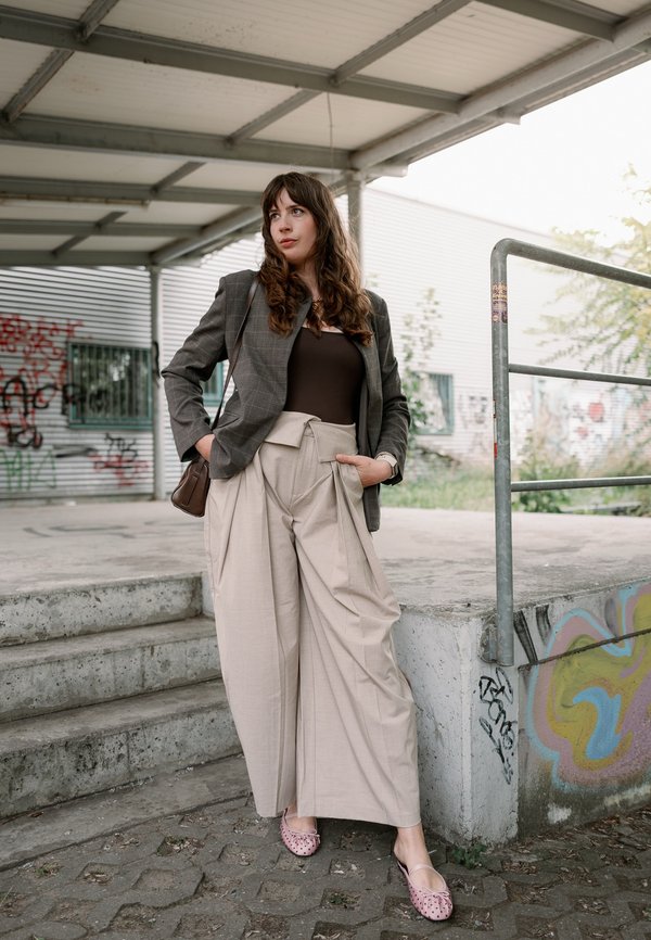 Young woman with long hair wearing beige wide pants, dark top, gray blazer, and pink flats, standing near concrete steps and graffiti wall outdoors.