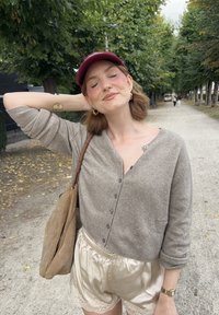 Cardigan gris avec un devant boutonné, associé à un short en satin clair avec des bordures en dentelle. Sac à épaule en daim marron, casquette bordeaux. Cadre décontracté en extérieur.