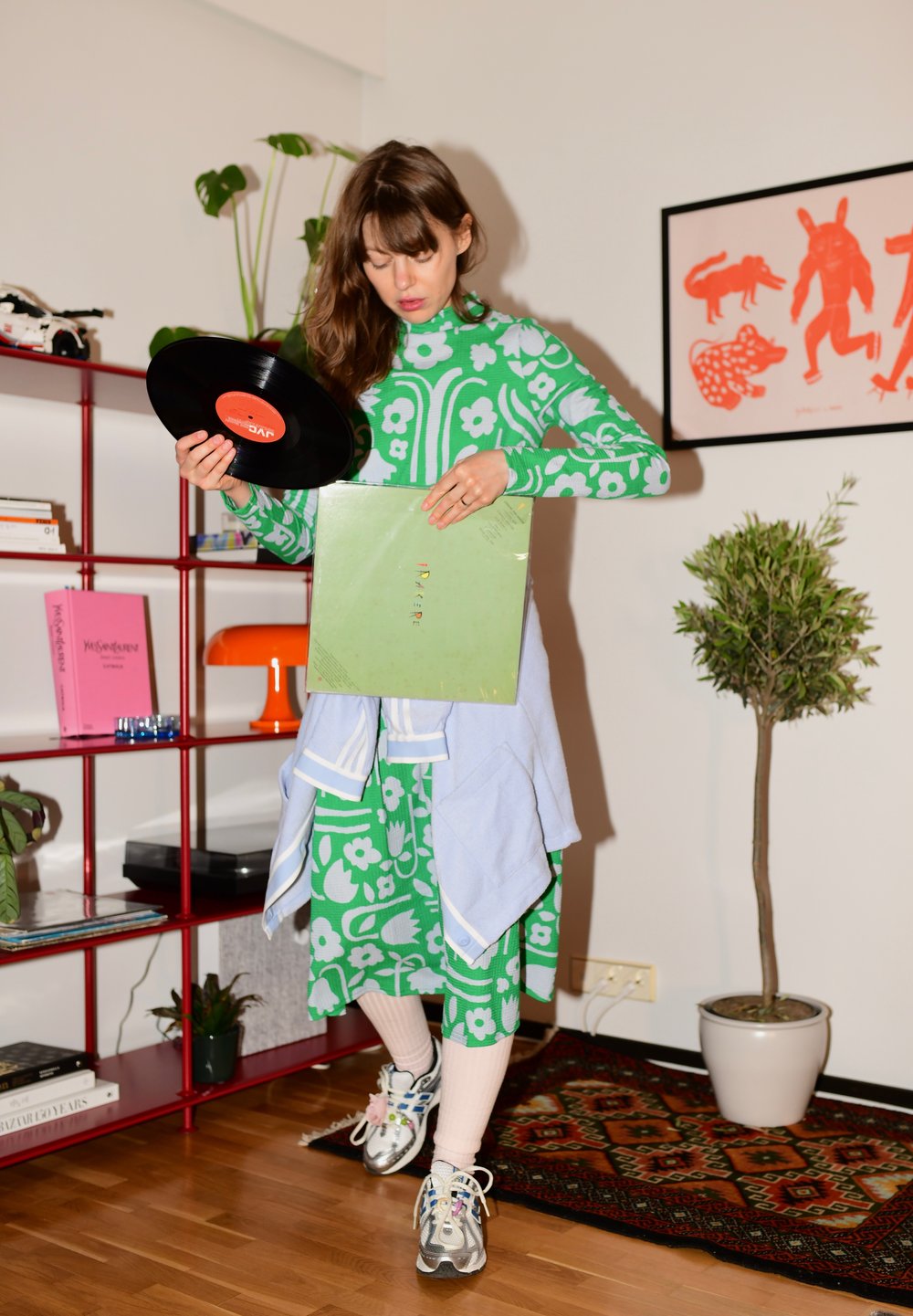 Modelo a usar um vestido verde com padrão, segurando um vinil preto e uma capa de disco verde. Ela tem um suéter azul claro amarrado à cintura.