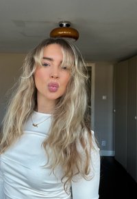 Jeune femme aux longs cheveux blonds ondulés portant un haut blanc, faisant un bisou, debout dans une pièce avec des portes fermées et une lumière au plafond.