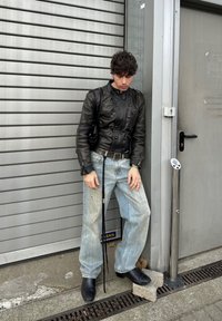 Ung person lænende mod en metalvæg og grå dør iført en sort læderjakke, lyseblå vide jeans og sorte støvler.