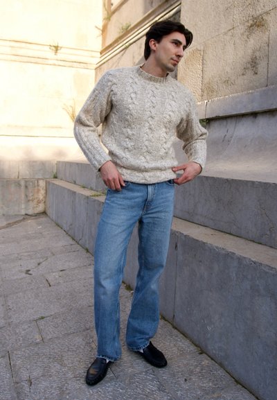 Joven con suéter beige de punto trenzado, jeans azules y zapatos negros, de pie al aire libre cerca de unos escalones de piedra.