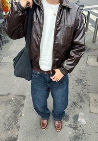 Personne portant une veste en cuir brun, un pull blanc, un jean bleu et des chaussures brunes, debout sur un trottoir de la ville tenant la sangle d'un sac noir.