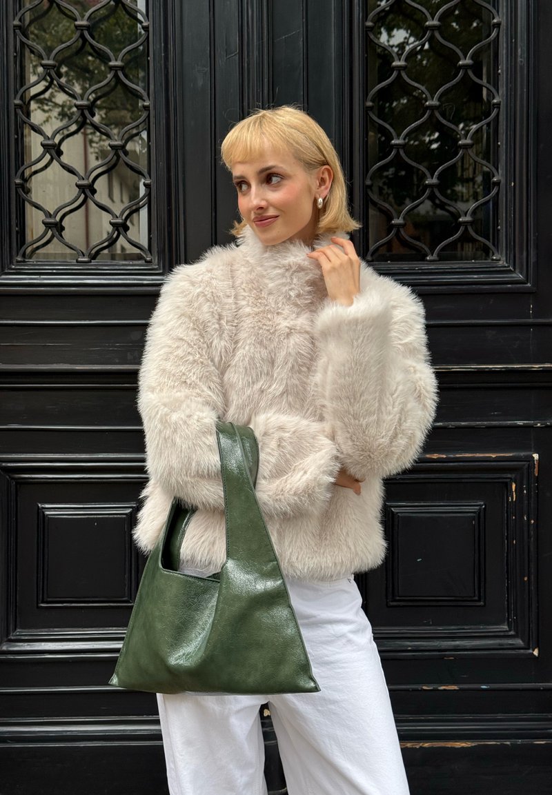 Donna bionda con una giacca pelosa beige chiaro e pantaloni bianchi tiene in mano una grande borsa di pelle verde scuro, in piedi davanti a una porta nera ornata.