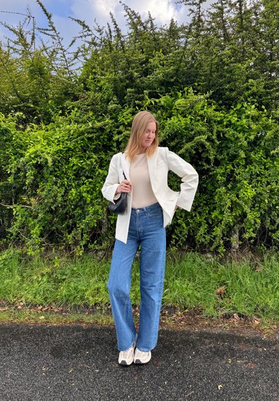 Mujer joven de pie sobre la acera, vestida con vaqueros azules, blazer blanco y zapatillas, sosteniendo un bolso negro frente a un fondo de arbustos verdes.