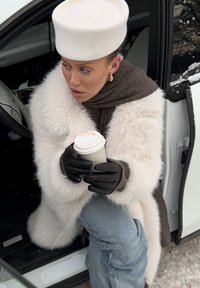 Femme portant un chapeau blanc, un manteau duveteux, des gants noirs et une écharpe, tenant une tasse de café tout en étant assise dans une voiture avec la portière ouverte par une journée enneigée.