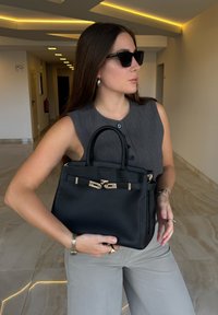 Černá kožená kabelka se strukturovaným designem, zlatými kovovými detaily a dvěma horními uchy. Šedý přizpůsobený bezrukávový top a světle šedé kalhoty.