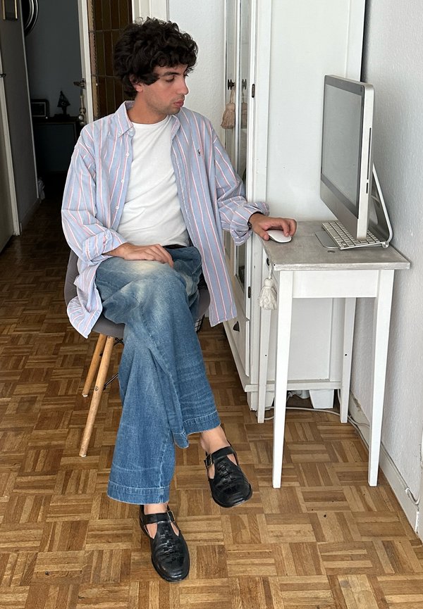 Hombre con cabello rizado y camisa a rayas sentado en una silla, usando un ratón de ordenador en un pequeño escritorio blanco con un monitor de sobremesa.