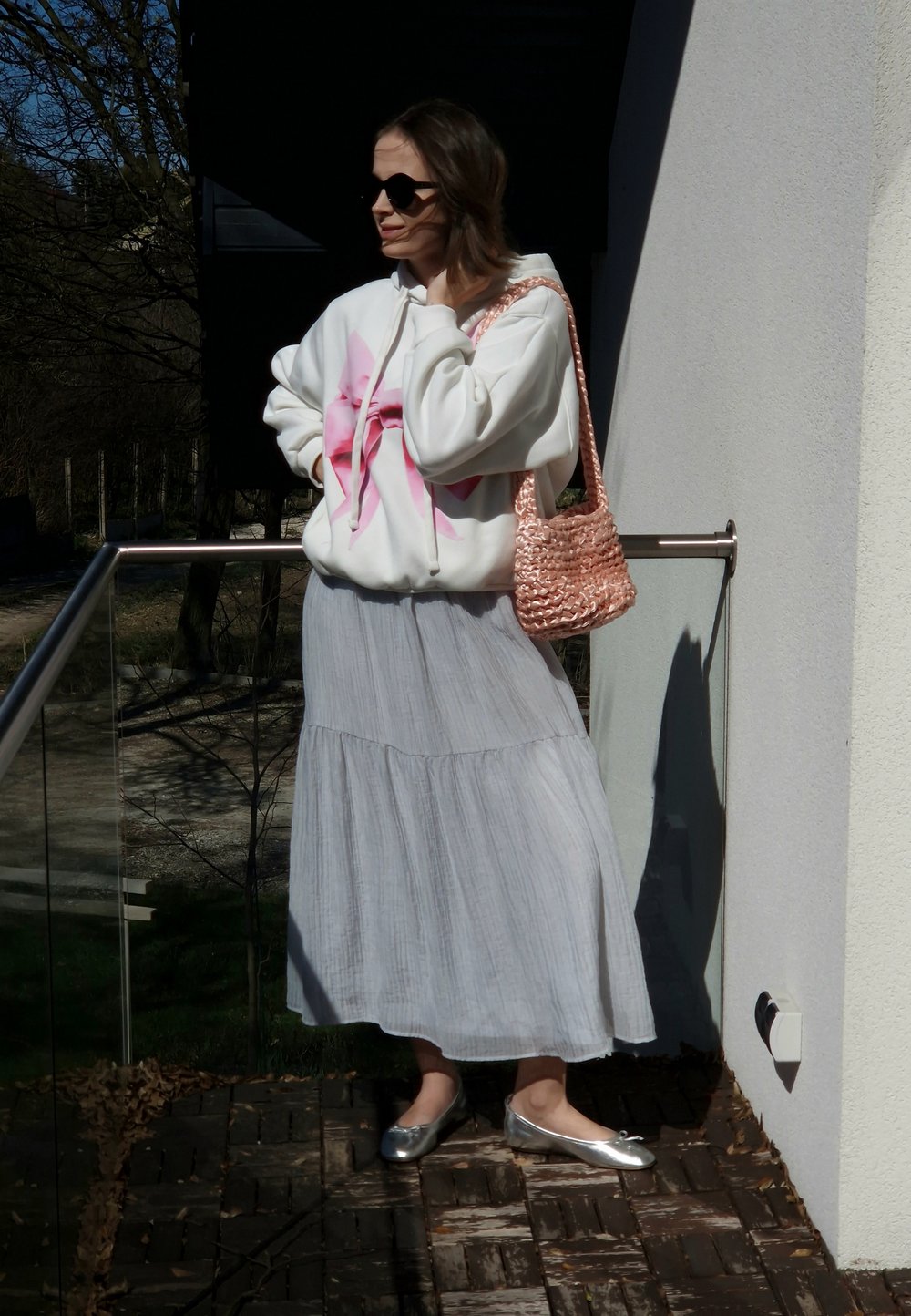 Bela kapuca z rožnatim tie-dye dizajnom, svetlo siva krilna krilka, srebrne balerinke in pletena torba v rožnato zlatem tonu. Na prostem.