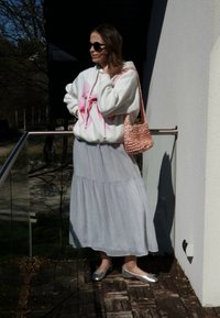 Bela kapuca z rožnatim tie-dye dizajnom, svetlo siva krilna krilka, srebrne balerinke in pletena torba v rožnato zlatem tonu. Na prostem.