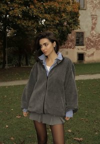 Mladá žena s tmavými vlasmi má na sebe šedú fleecovú bundu cez modrú károvanú košeľu a šedú sukňu, stojí na tráve blízko jesenných stromov a starej budovy.