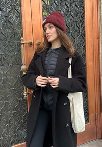 Cappotto nero con grandi rever sovrapposto a una giacca nera imbottita, abbinato a un berretto di maglia bordeaux e una borsa a tracolla crema davanti a una porta ornata.