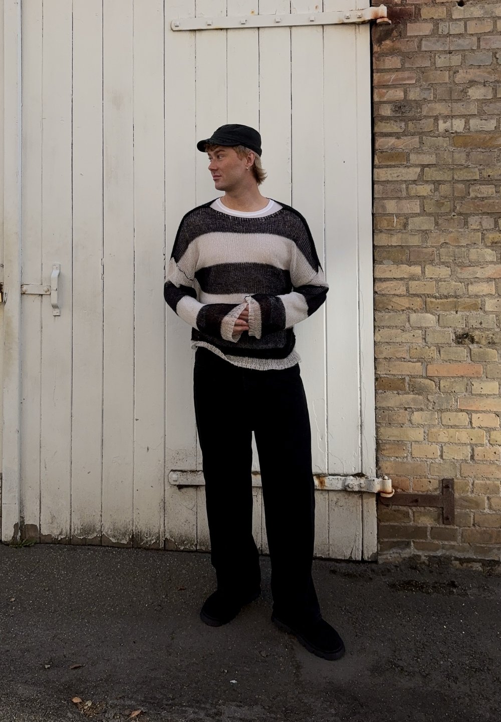 Oversized, gestreept sweater in zwart en crème met een brede halslijn en lange mouwen, gecombineerd met zwarte broeken en een zwarte pet.