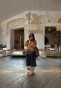 Casaco de camurça castanho sobre uma camisa castanha, calções largos pretos e botas de fivela em tom de areia. Segura uma bebida, de pé numa sala com espelhos e decoração vintage.