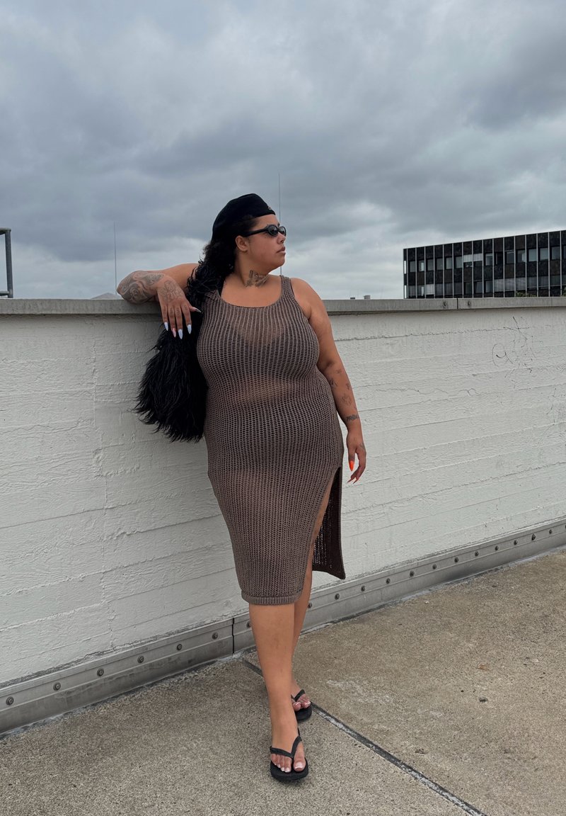 Vestido midi castanho de malha com uma fenda lateral, design sem mangas, combinado com chinelos pretos e estilizado com cabelo longo e preto e unhas.