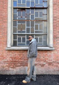 Uomo con occhiali da sole, giacca grigia e pantaloni beige in piedi di lato contro un muro di mattoni con una grande finestra vintage con telaio in metallo.