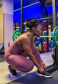 Femme en tenue de sport violette attachant ses chaussures près d'un rack de poids avec des disques de musculation colorés dans une salle de gym éclairée en bleu.