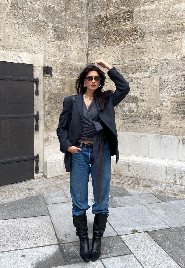 Blazer oversize a righe nere sopra un top grigio avvolgente, jeans loose di lavaggio medio e stivali neri con tacco al ginocchio. Accessoriato con occhiali da sole.