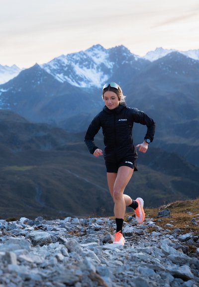 Atleta vestido con una chaqueta aislante negra y pantalones cortos negros, corriendo sobre terreno rocoso con montañas al fondo, zapatillas de correr naranjas.