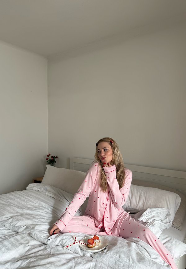 Joven mujer en pijama rosa con estampado de corazones sentada en una cama blanca, con un postre de fresa en un plato cercano, en un dormitorio minimalista.