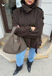 Femme portant un manteau croisé marron foncé, un jean bleu clair, des bottes pointues noires, tenant un grand sac à main tressé marron, debout en plein air.