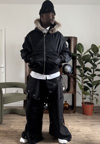 Veste bomber noire avec capuche doublée de fourrure, associée à un pantalon noir oversize orné de détails œillets et une chemise blanche en dessous. Cadre intérieur.