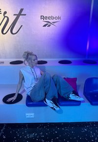 Jeune femme portant un sweat à capuche sans manches et un pantalon ample, assise sur un banc bleu sous un éclairage violet, chaussée de baskets Reebok blanches et noires.