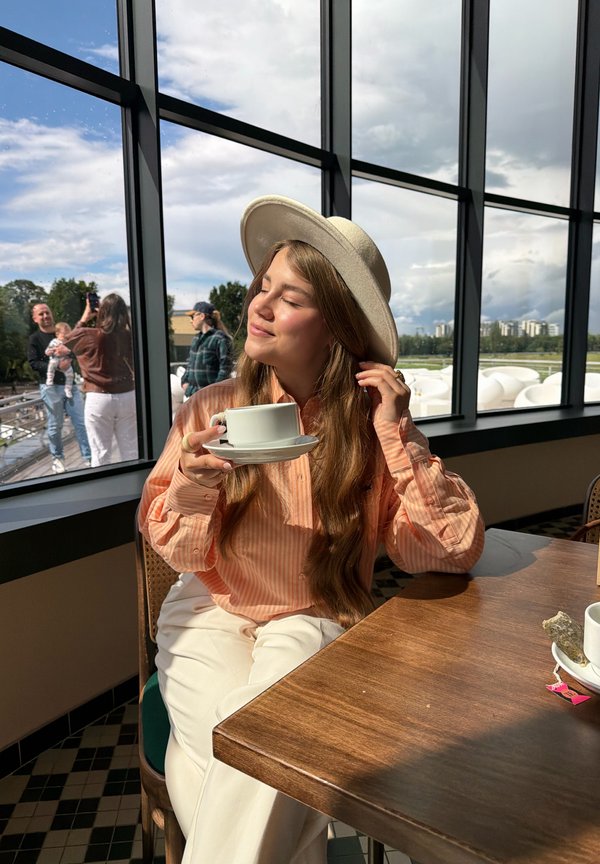 Young woman in a hat enjoying a cup of tea by a large window with a bright outdoor view and people in the background.