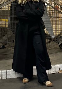 Long manteau noir par-dessus un sweat à capuche noir et un pantalon, assortis à des chaussures beiges et marron. Le décor présente un fond en grille métallique.
