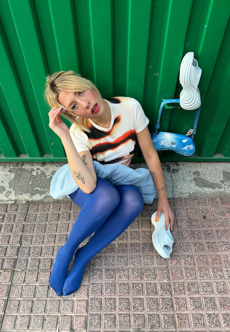 Femme aux cheveux blonds assise sur le trottoir contre un mur vert, portant un t-shirt graphique, des collants bleus, tenant des sandales blanches sculpturales.
