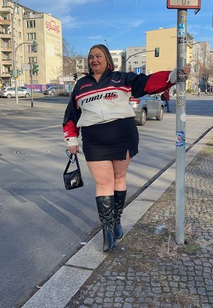 Chaqueta FUBU negra y roja sobre una falda corta negra, combinada con botas negras hasta la rodilla. Sosteniendo un pequeño bolso negro, de pie junto a un poste en la calle.