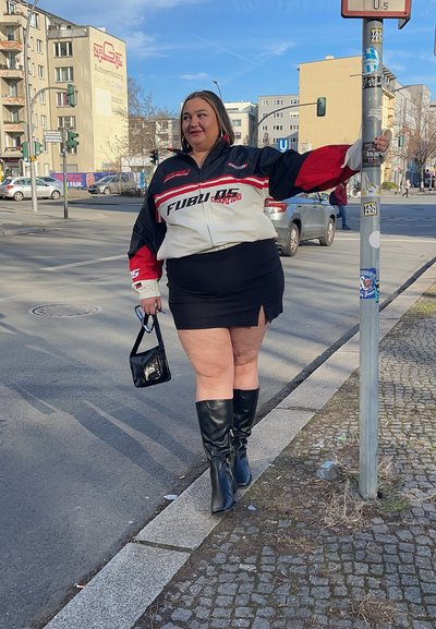 Chaqueta FUBU negra y roja sobre una falda corta negra, combinada con botas negras hasta la rodilla. Sosteniendo un pequeño bolso negro, de pie junto a un poste en la calle.