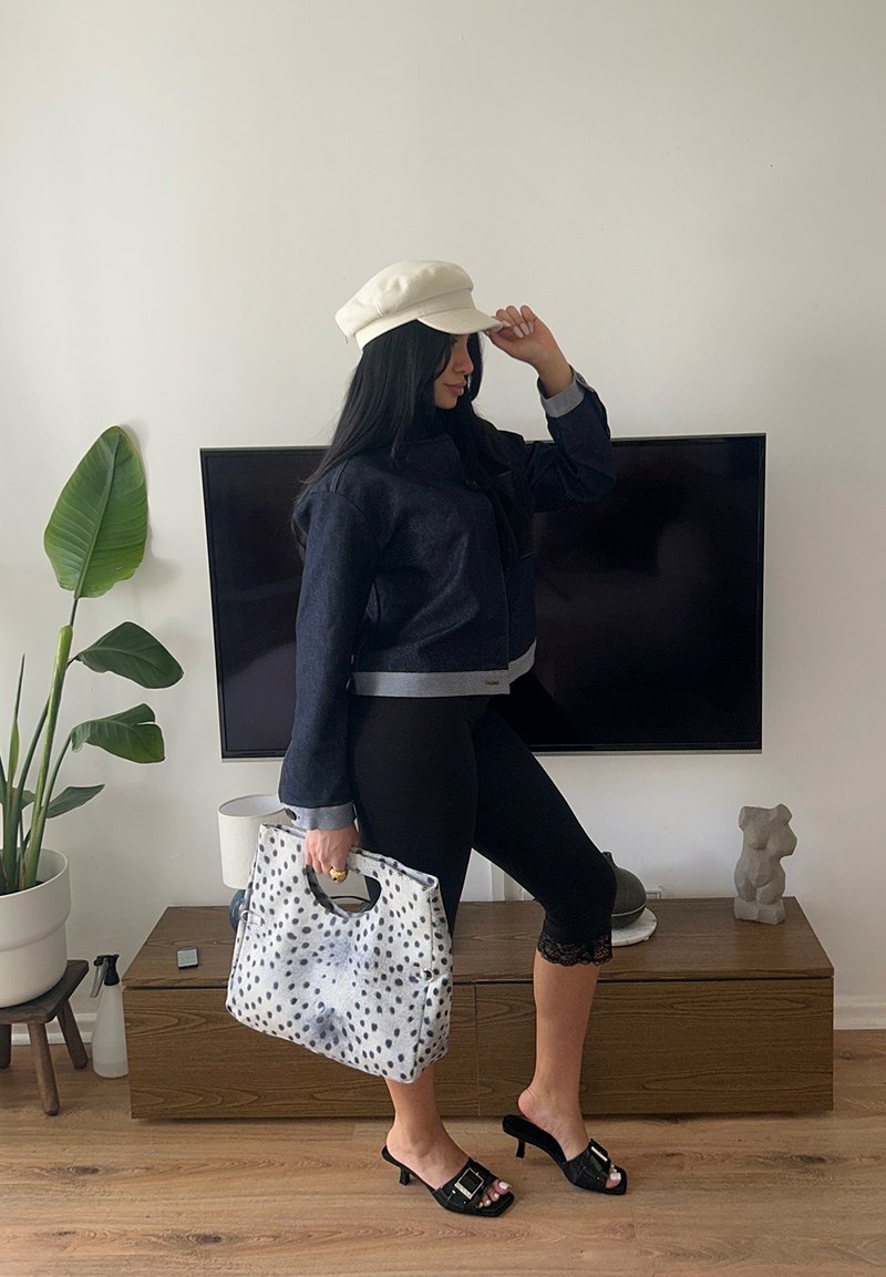 Femme en veste noire, leggings, casquette beige et talons noirs, tenant un sac à main blanc à taches noires, debout dans un salon avec une télévision et une plante.