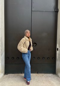 Krämfärgad, fluffig jacka över en svart topp, parat med högmidjade blå utsvängda jeans och vinröda sneakers, mot en mörk dörr.