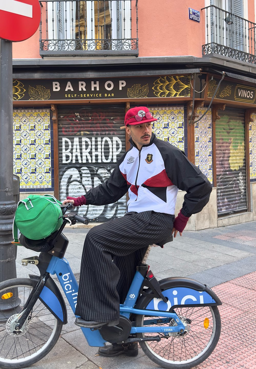 Uma bicicleta azul com um banco preto. Uma pessoa usa um boné vermelho, uma jaqueta branca e preta, e calças listradas, segurando uma bolsa verde na bicicleta.
