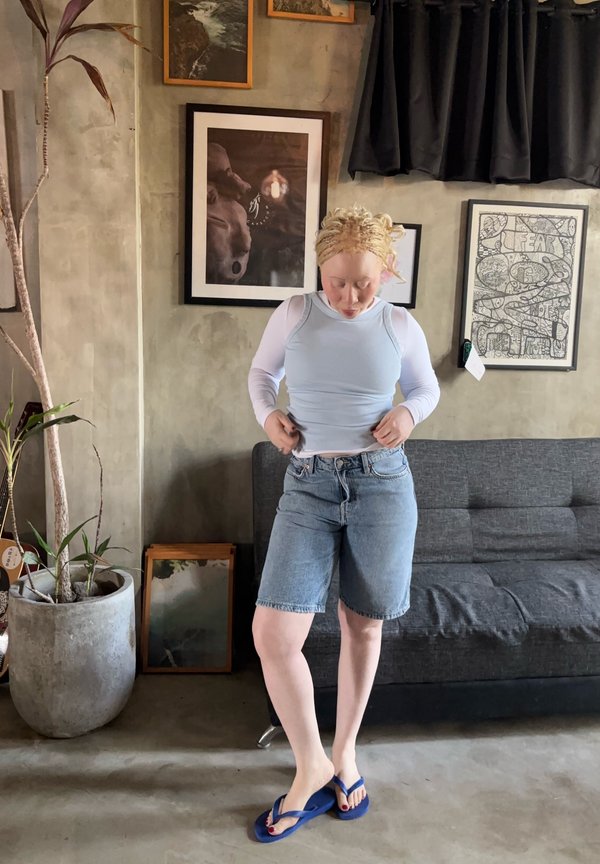 Person mit heller Haut und blondem Haar in Jeansshorts und weißem Oberteil richtet ihre Kleidung in einem Wohnzimmer mit grauem Sofa und gerahmter Kunst.