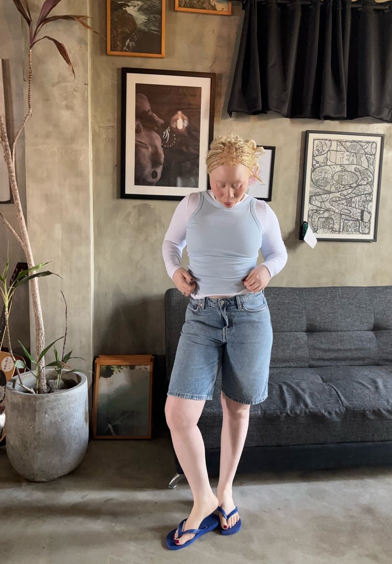 Personne à la peau claire et aux cheveux blonds en short en jean et haut blanc ajustant ses vêtements dans un salon avec un canapé gris et des œuvres d'art encadrées.