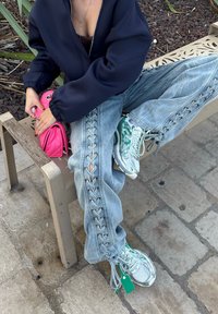 Osoba sjedi na bež metalnoj klupi, obučena u plave traperice s vezanjem, zelene i bijele tenisice, mornarsko plavu jaknu i drži ružičastu pamučnu torbu.