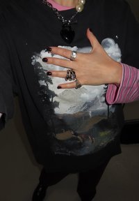 Main avec vernis à ongles foncé portant des bagues en argent sur un t-shirt noir avec un imprimé de montagne et forêt, et une manche superposée rayée rose.